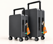 Two dark gray hard-shell spinner suitcases with telescoping handles, shown on a clean white studio background. The larger suitcase stands behind the smaller one, and both have bright orange luggage tags attached.