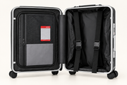An open hard-shell suitcase with a silver frame and black interior, showing a zippered compartment with a mesh pocket and a red-and-white luggage tag on the left, plus two black compression straps on the right, photographed on a clean white background.